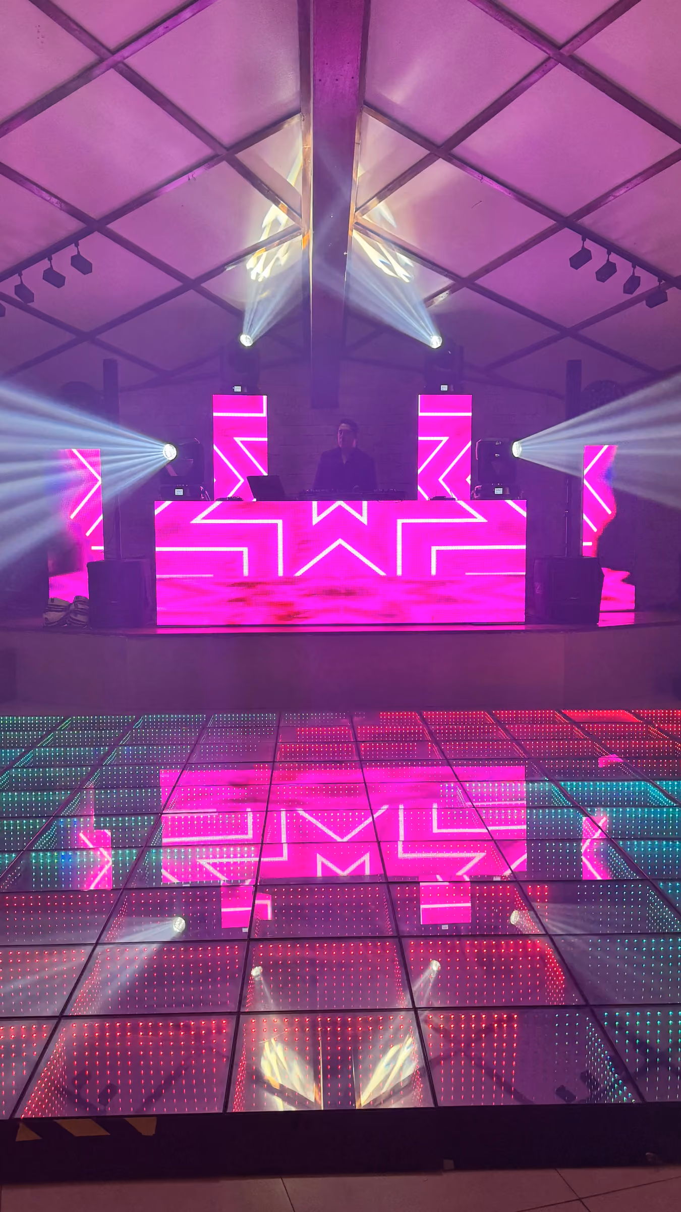 Cabina de DJ con pantallas LED rosadas, luces robóticas y piso iluminado