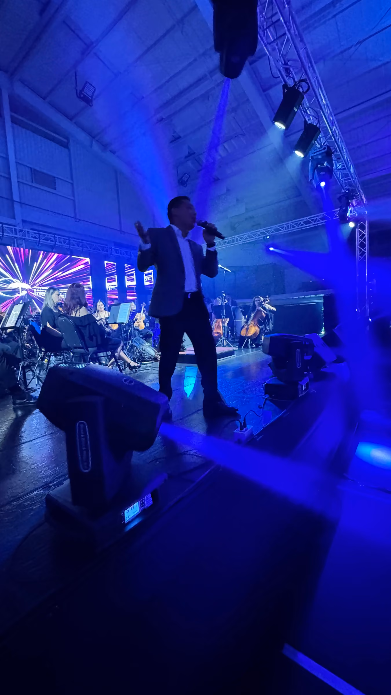 Cantante y orquesta en escenario bajo luces azules y truss